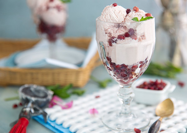 Pomegranate Semifreddo in a tall clear glass with an ice cream scoop in the background on a white ceramic serving trivet