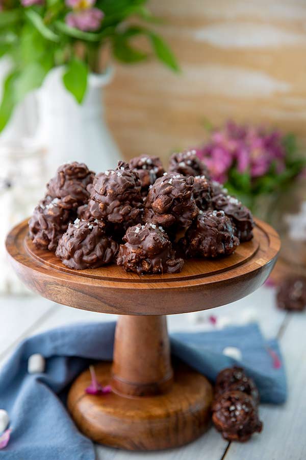 Chocolate Sunbutter Cookie Clusters on a wooden pedestal with purple flowers in vases in the background