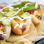 Nacho-inspired hot dogs on a bed of tortilla chips with a drizzle of sour cream and topped with sliced avocado