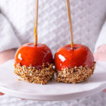 Person in a white sweater holding out a white tray with Caramel Apples dipped in crushed hazelnuts