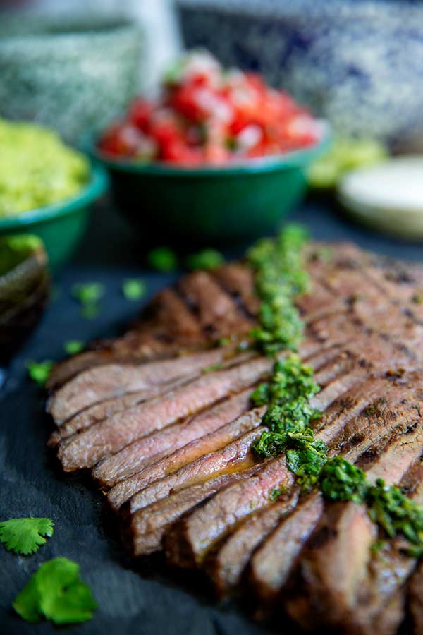 Carne Asada sliced into strips with chimichurri drizzled on top and fresh pico de gallo in the background