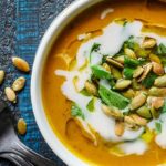Closeup of Paleo Curried Pumpkin Soup in a white bowl on a dark blue background topped with pumpkin seeds and coconut cream