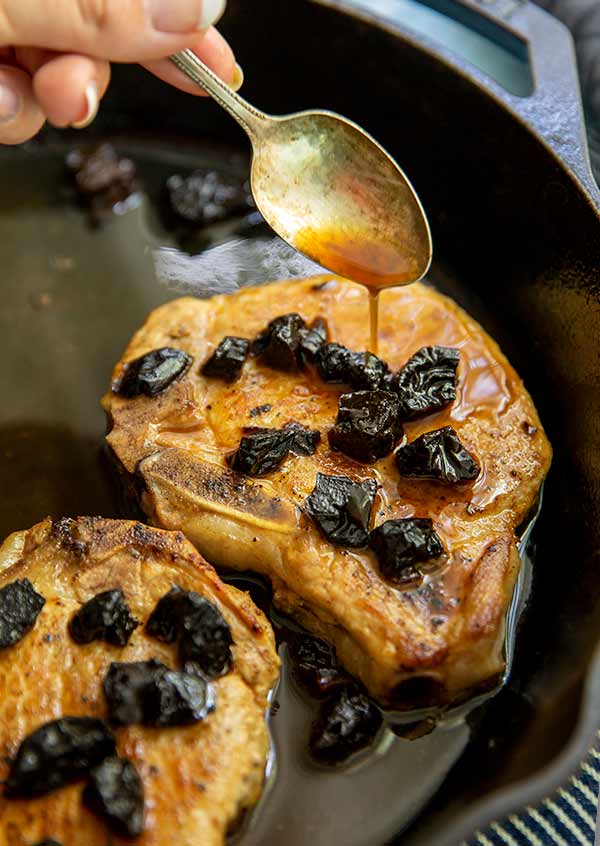 Plum-Glazed Pork Chops in a cast iron skillet with a woman's hand spooning glaze over top