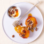 Roasted Peaches on a white plate with walnuts and honey on top and a wooden honey wand next to it