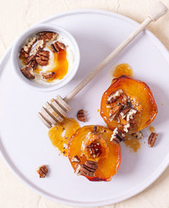 Roasted Peaches on a white plate with walnuts and honey on top and a wooden honey wand next to it
