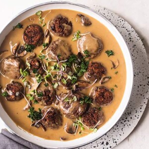 Vegan Swedish Meatballs in gravy in a white dish with a white and black speckled plate underneath