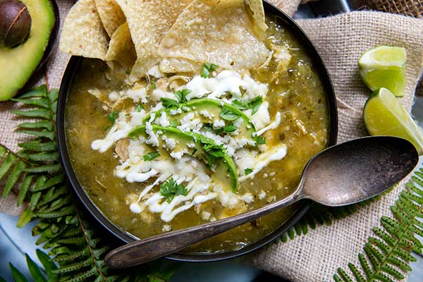 Closeup of Tomatillo Soup Recipe in a black bowl with tortilla chips, crema, and avocado on top