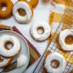 Overhead view of frosted Pumpkin Spice Donuts on a plate and metal cooling rack on top of yellow plaid tablecloth