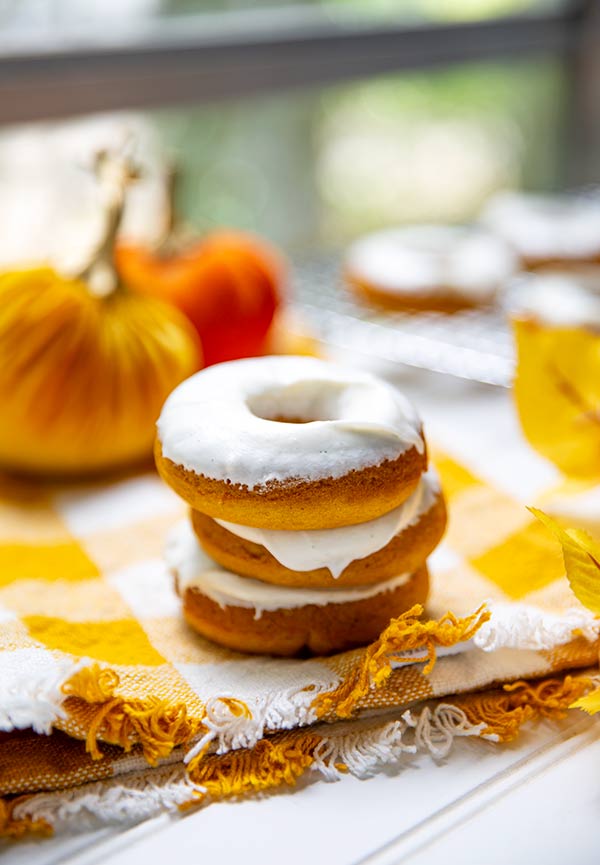 Stack of three Pumpkin Spice Donuts on a yellow plaid tablecloth with fall leaves and pumpkin statues in the background