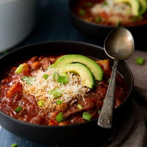 Vegetarian Chili topped with avocado slices and green onions in a black bowl on a dark blue moody background