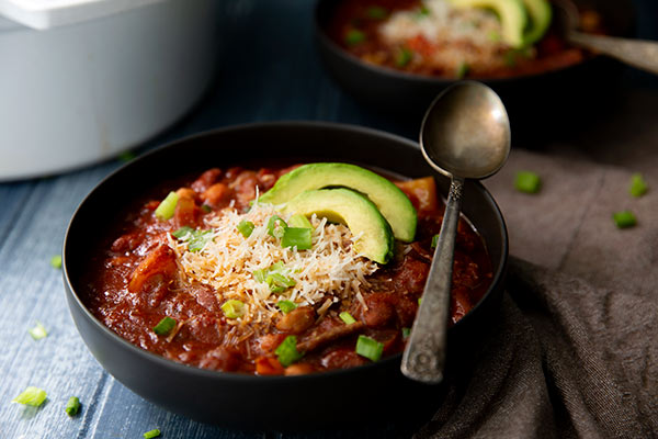 Vegetarian Chili topped with avocado slices and green onions in a black bowl on a dark blue moody background
