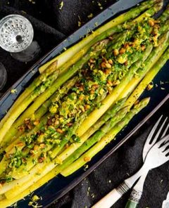 Asparagus Gremolata on a black oval serving tray on top of a black tablecloth with crystal glasses around it