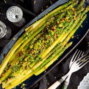 Asparagus Gremolata on a black oval serving tray on top of a black tablecloth with crystal glasses around it