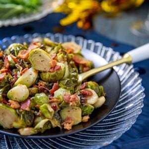 Bacon and Maple Roasted Brussels Sprouts on a black plate set over a clear serving plate on a navy blue tablecloth