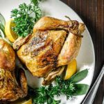 Overhead view of Cornish Game Hens on a white plate with fresh herbs and lemon slices surrounding them