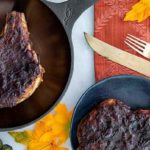 Overhead view of Cranberry Sauce Pork Chops on a black cast iron skillet and black plate on an orange tablecloth with fall decor