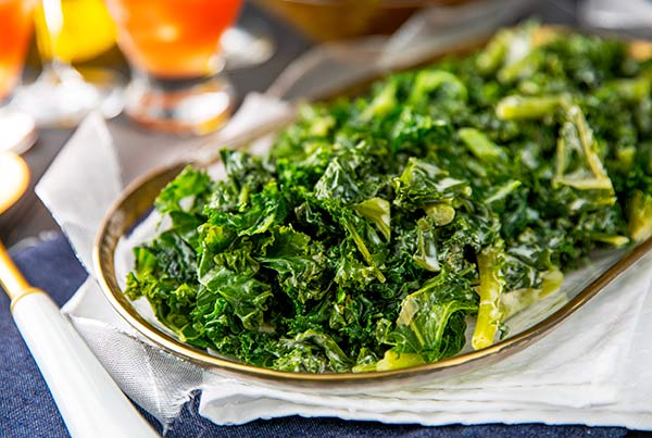 Closeup of Creamed Kale on a clear oval serving tray with white cloth napkins underneath on a dark blue background