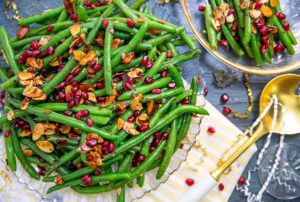 Overhead view of Holiday Green Beans with pomegranates and roasted almonds in clear glass dishes with a gold serving spoon next to it