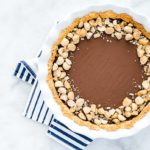 Macadamia Nut Chocolate Pie with Coconut Crust in a white ceramic pie pan on a white marble background with a navy and white striped napkin underneath