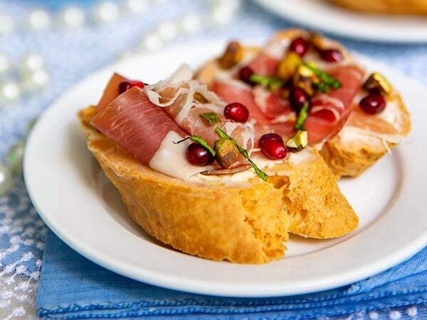 Closeup of Prosciutto, Pomegranate & Pistachio Bruschetta on a white plate with blue napkins and lace tablecloth