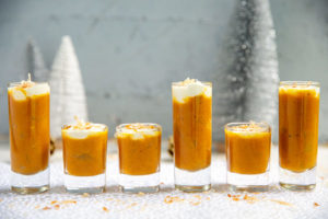 Pumpkin Apple Soup Shooters with Coconut Cream lined up in different sized shot glasses on a silver and white holiday background