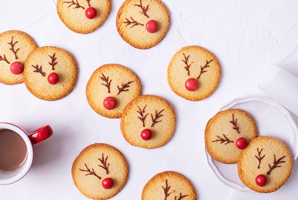 Rudolph Cookies scattered across a white background with a red mug of hot cocoa