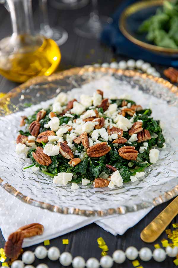 Sauteed Spinach with Feta and Walnuts on a clear glass plate lined with gold on a dark background with pearls and gold confetti