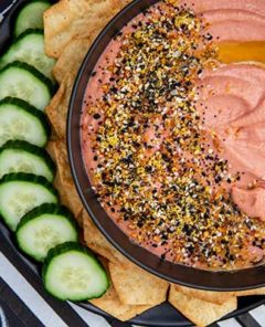 Overhead view of Beet Hummus with everything bagel seasoning on top and sliced cucumber and gluten-free crackers on a white and black striped background with football themed decor