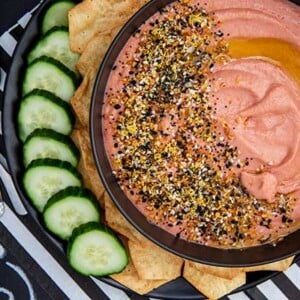 Overhead view of Beet Hummus with everything bagel seasoning on top and sliced cucumber and gluten-free crackers on a white and black striped background with football themed decor