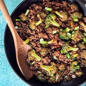 Overhead view of ground bison and broccoli skillet dinner in a black skillet with wooden spoon on a sky blue background