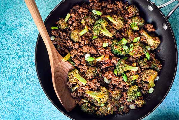 Overhead view of ground bison and broccoli skillet dinner in a black skillet with wooden spoon on a sky blue background