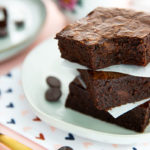Closeup of Fudgy Brownies stacked on a white plate with light pink background and gold fork and napkin with hearts on it