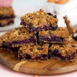 Granola-Topped Blueberry Pie Bars stacked on a round wooden cutting board on a light pink background