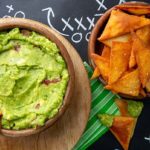 Keto Chips and Guacamole in wooden bowls on a black chalkboard with football game plays written in chalk