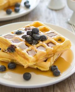 Lemon Blueberry Waffles on a white plate topped with syrup and fresh blueberries on a light wood table