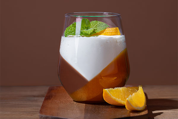 No-Bake Chocolate Pumpkin Dessert in a stemless wine glass set on a dark wood cutting board with a dramatic brown background and orange slices to garnish