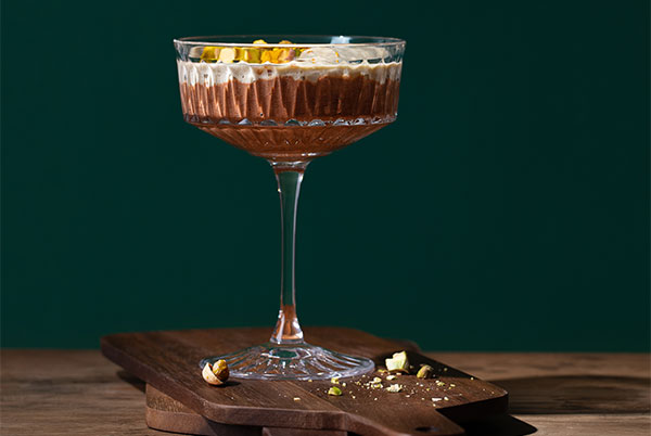 Pistachio Chocolate Mousse in a fancy cocktail glass set on a dark wood cutting board with a dramatic dark green background