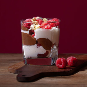 Raspberry Chocolate Panna Cotta in a square glass on a dark wood cutting board with a dark maroon background