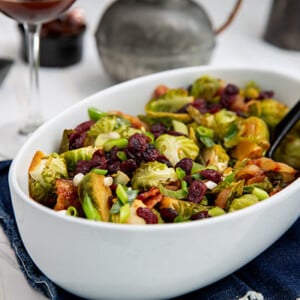 Soy-Glazed Brussels Sprouts in a white oval serving dish with glasses of red wine in the background