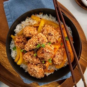 Overhead view of Chinese Salt & Pepper Chicken in a black bowl set on top of a blue napkin on a wooden serving board with wooden chopsticks