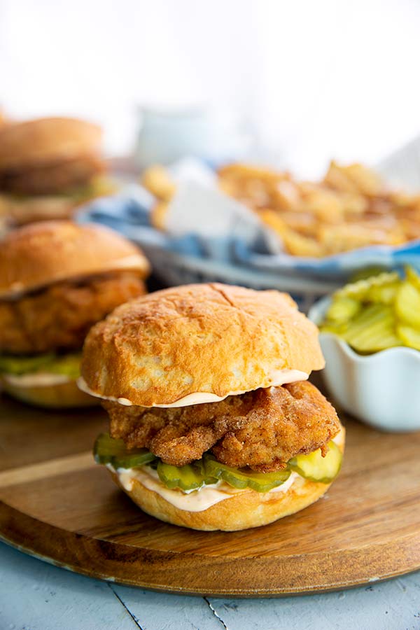 Fried Chicken Sandwich on a wooden board with a white bowl of pickles and basket of gravy fries in the background