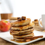 Stack of Granola Pancakes on a square white plate with apples in the background