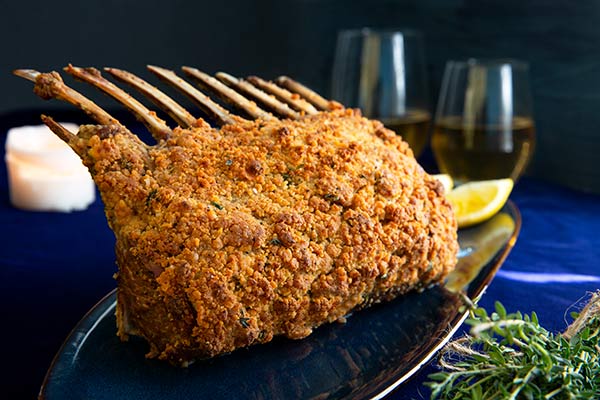 Panko and Goat Cheese Crusted Rack of Lamb on a dark blue plate on a dark blue tablecloth with two glasses of white wine in the background