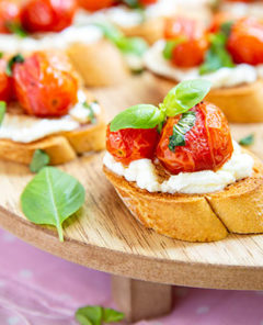 Roasted Tomato and Ricotta Basil Crostini spread around a raised wooden serving board on a light pink tablecloth