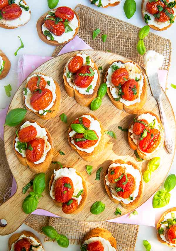Overhead view of Roasted Tomato and Ricotta Basil Crostini scattered on and around a round wooden cutting board with fresh basil scattered around