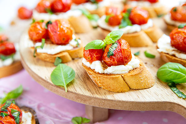 Roasted Tomato and Ricotta Basil Crostini spread around a raised wooden serving board on a light pink tablecloth