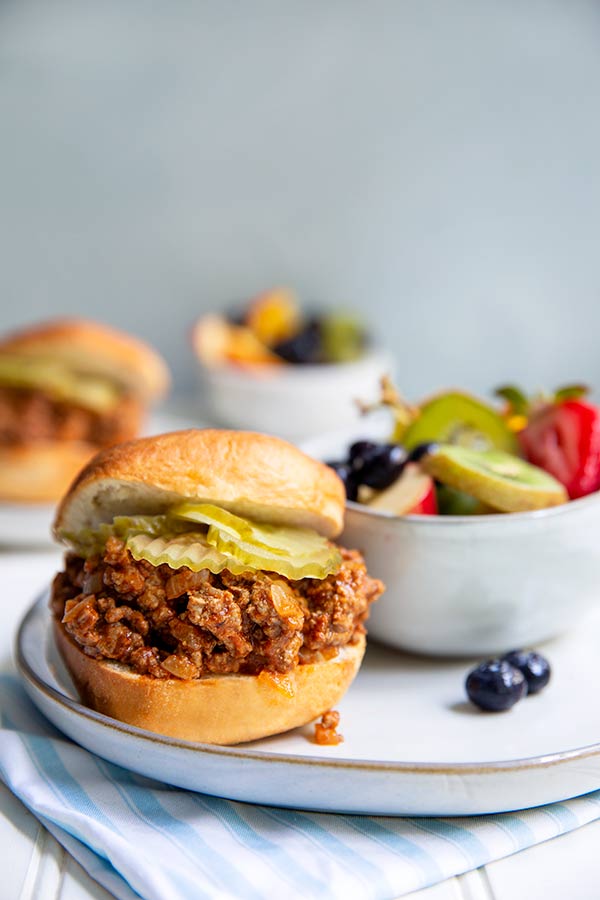Sloppy Joes on a white plate with a bowl of fresh sliced fruit in the background