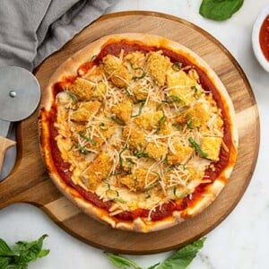 Overhead view of Chicken Parm Pizza on a wooden pizza board with pizza cutter on the size and basil leaves around the board
