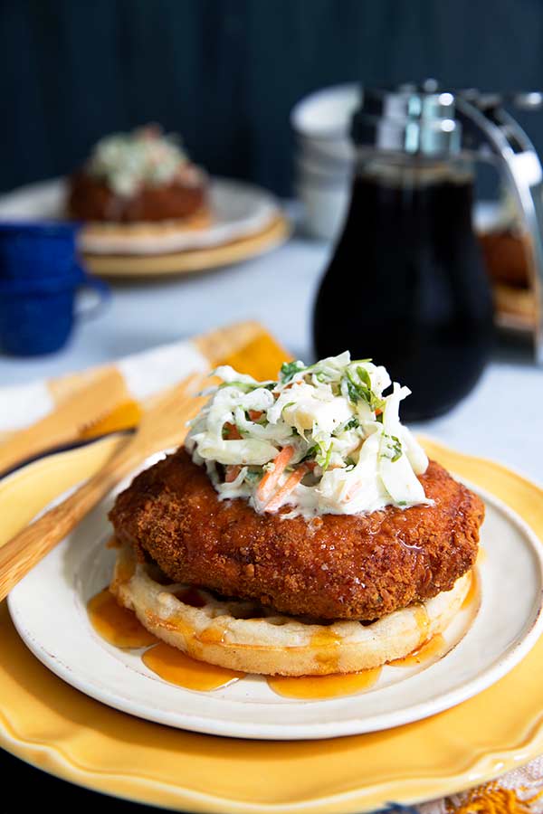 Gluten-free Chicken and Waffles topped with coleslaw on a white plate over a yellow plate and yellow napkin