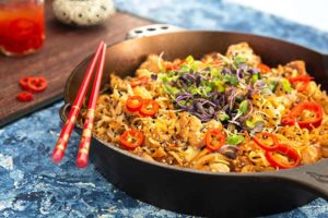 Closeup of Spicy Citrus Chicken and Noodles topped with red peppers in a skillet on a blue background with red chopsticks